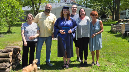 Scott with Cali in her cap and gown holding her diploma, with four family members on a sunny lawn