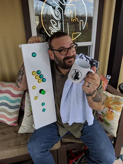 Scott on a porch holding a Northwoods Computer Guy coffee mug and a white board with colorful buttons